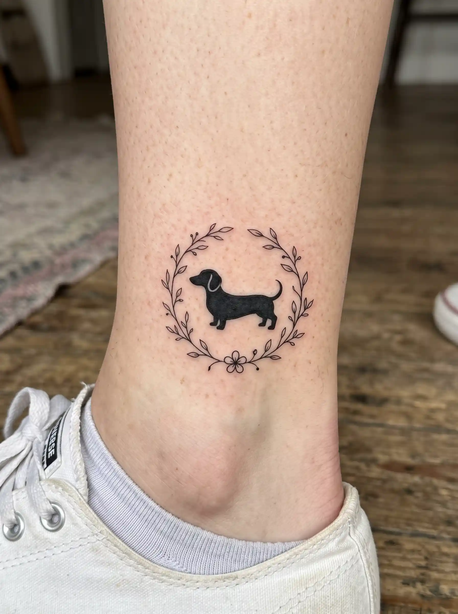 Minimalist black dachshund silhouette tattoo in floral wreath on ankle